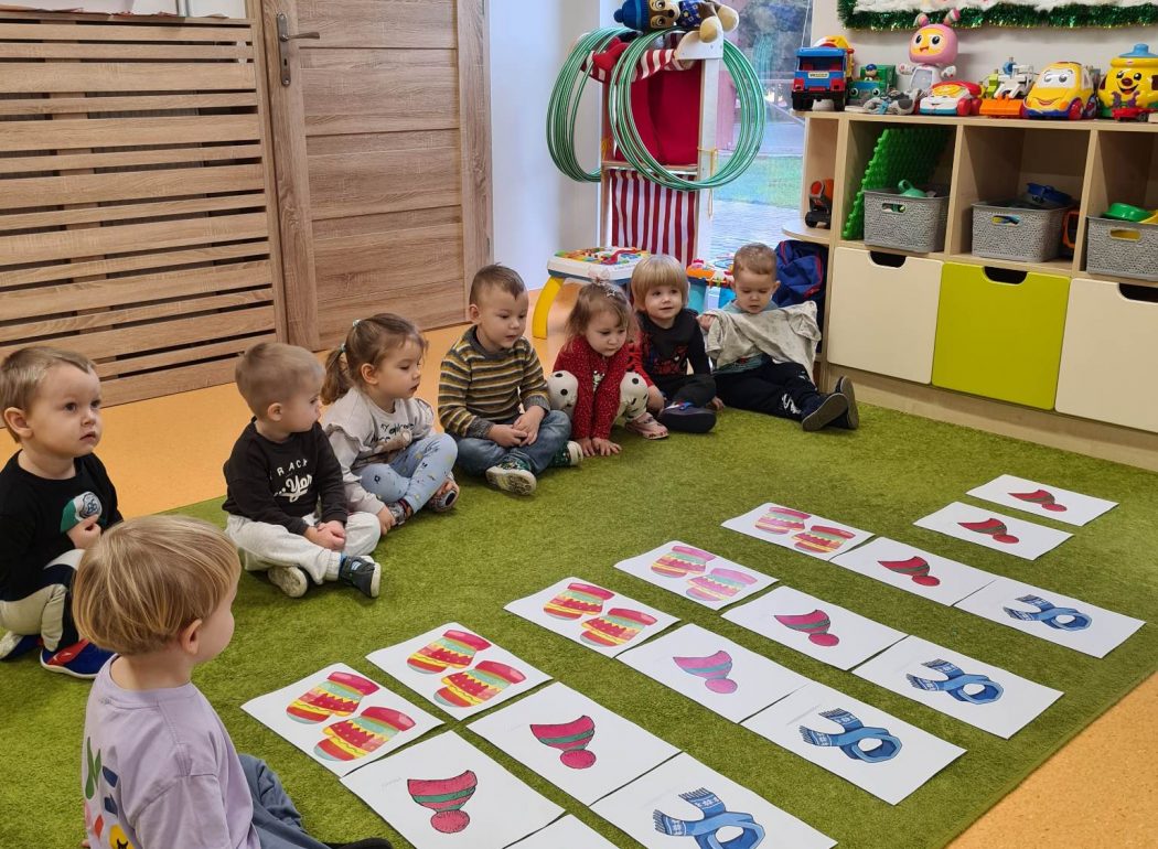 Dzieci siedzą na dywanie koło siebie. Przed dziećmi rozłożone są ilustracje związane z zimową garderobą.