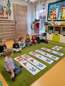 Dzieci siedzą na dywanie koło siebie. Przed dziećmi rozłożone są ilustracje związane z zimową garderobą.