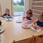 Dzieci siedzą przy stoliku ubrane w niebieskie fartuszki i malują farbami kontury rękawiczek.