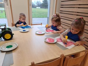 Dzieci siedzą przy stoliku ubrane w niebieskie fartuszki i malują farbami kontury rękawiczek.