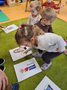 Dziewczynka pokazuje na obrazku Babcie i Dziadka . W tle dzieci oglądają obrazki z członkami rodziny.