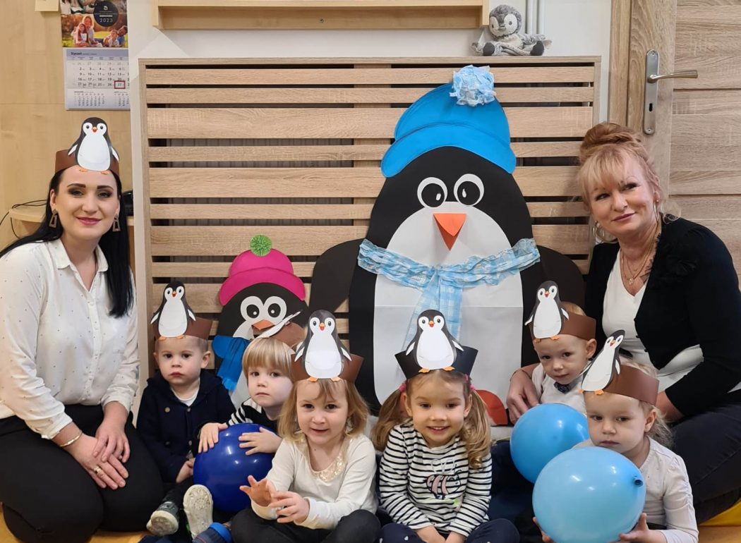 Dzieci wraz z opiekunkami ubrani na biało-czarno z opaskami pingwina siedzą koło siebie pozując do zdjęcia grupowego.