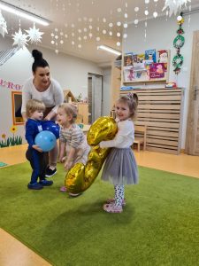Opiekunka wraz z dziećmi stoi na zielonym dywanie trzymając kolorowe baloniki.