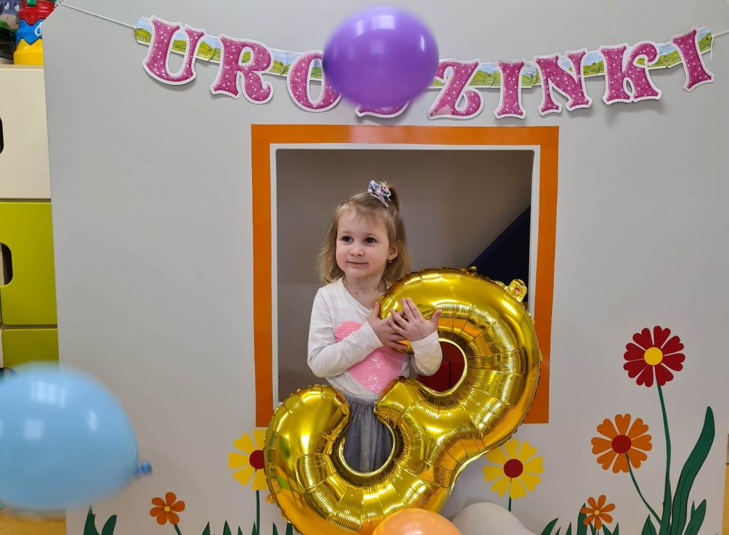 Dziewczynka stoi i trzyma duży złoty balon w kształcie cyfry trzy. W tle widać urodzinowy napis.