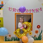 Dziewczynka stoi i trzyma duży złoty balon w kształcie cyfry trzy. W tle widać urodzinowy napis.