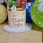 Na zdjęciu widać tort urodzinowy z postaciami z bajek. Koło tortu leżą kolorowe baloniki.