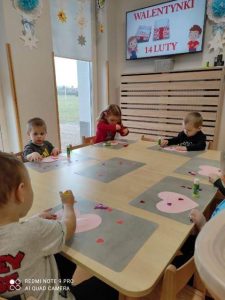 Dzieci siedzą przy stoliku i wyklejają różowe serduszko, czerwonymi serduszkami tworząc z nich piękną walentynkę.