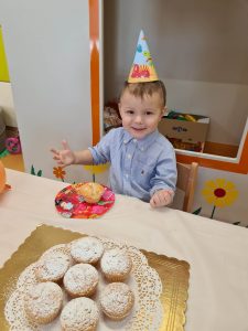 Chłopiec siedzi przy stoliku ubrany w urodzinową czapkę i częstuje się muffinkami.