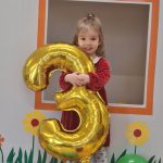 Dziewczynka stoi i trzyma duży balon w kształcie cyfry trzy.