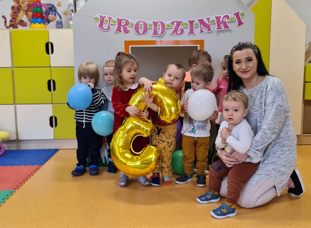 Dzieci stoją i pozują do zdjęcia z solenizantką która trzyma duży złoty balon w kształcie cyfry trzy.