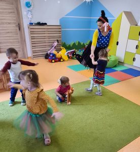 Na zielonym dywanie dzieci ubrane na kolorowo tańczą do piosenek. W tle widać sale zabaw.