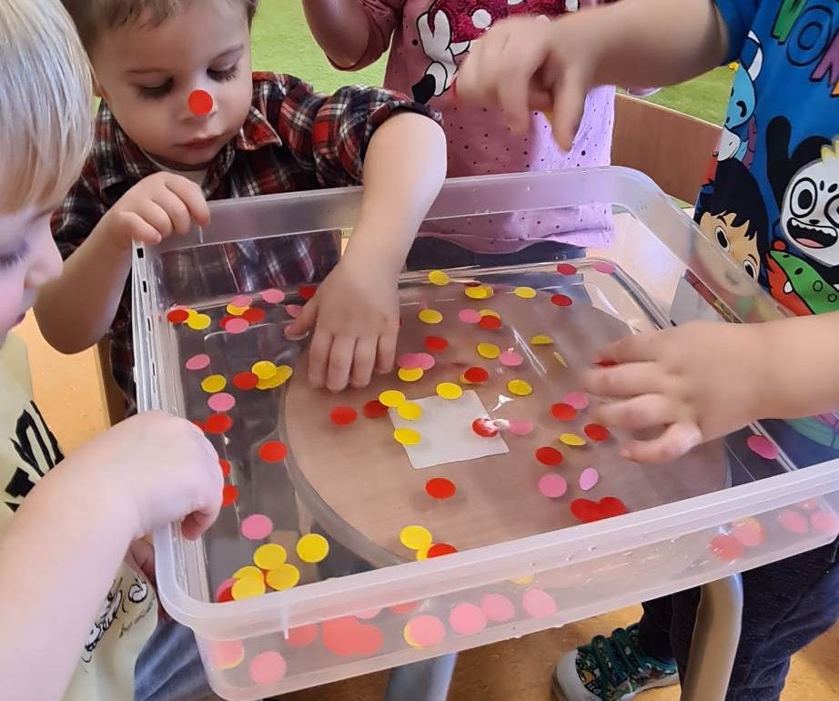 Dzieci stoją koło siebie i z pojemnika wyciągają kolorowe kółeczka zamoczone w wodzie i naklejają je na buzi kolegi lub koleżanki