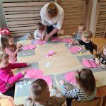 Dzieci siedzą przy stoliku i na różowej kartce przyklejają białe kwadraciki tworząc z nich jamę ustną.