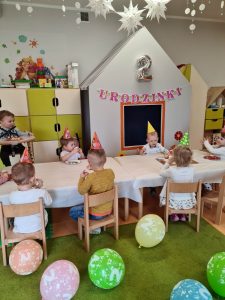 Dzieci siedzą przy stoliku w urodzinowych czapeczkach i częstują się babeczkami.