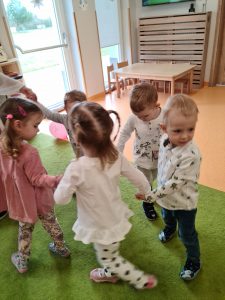 Dzieci trzymają się za ręce i tańczą w kółeczku do piosenek urodzinowych.
