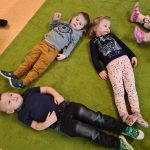Dzieci leżą na zielonym dywanie tworząc duży trójkąt.