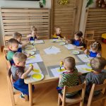 Dzieci siedzą przy stoliku ubrane w niebieskie fartuszki i malują na kartce żółtą farbą.