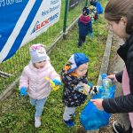 Dzieci ubrane w niebieskie rękawiczki zbierają śmieci na trawie i wrzucają do niebieskiego worka.