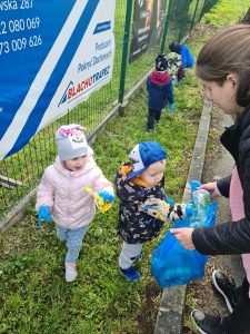 Dzieci ubrane w niebieskie rękawiczki zbierają śmieci na trawie i wrzucają do niebieskiego worka.
