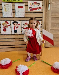 Dziewczynka siedzi na krzesełku i trzyma w rączkach flagę polski i kwiat biało - czerwony. W tle widać powieszone symbole narodowe.