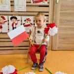 Chłopiec siedzi na krzesełku i trzyma w rączkach flagę polski i kwiat biało - czerwony. W tle widać powieszone symbole narodowe.