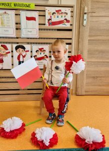 Chłopiec siedzi na krzesełku i trzyma w rączkach flagę polski i kwiat biało - czerwony. W tle widać powieszone symbole narodowe.