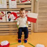 Chłopiec siedzi na krzesełku i trzyma w rączkach flagę polski i kwiat biało - czerwony. W tle widać powieszone symbole narodowe.