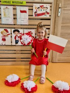 Dziewczynka siedzi na krzesełku i trzyma w rączkach flagę polski i kwiat biało - czerwony. W tle widać powieszone symbole narodowe.
