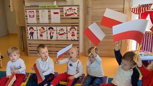 Dzieci siedzą na podusiach i do piosenki o Polsce machają flagami.