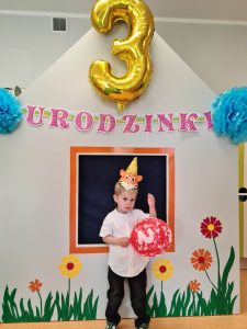 Solenizant stoi i pozuje do zdjęcia w rączkach trzyma czerwony balon. W tle widać napis urodzinki i duży złoty balon w kształcie cyfry trzy.