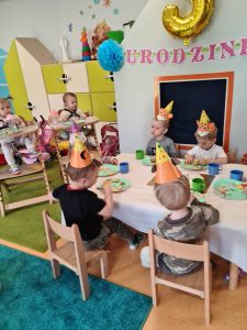Dzieci ubrane w urodzinowe czapeczki siedzą przy stoliku i częstują się urodzinowym torcikiem z babeczek.