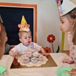Chłopiec ubrany w urodzinową czapeczkę dmucha świeczkę na urodzinowym torcie zrobionym z babeczek.