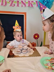 Chłopiec ubrany w urodzinową czapeczkę dmucha świeczkę na urodzinowym torcie zrobionym z babeczek.