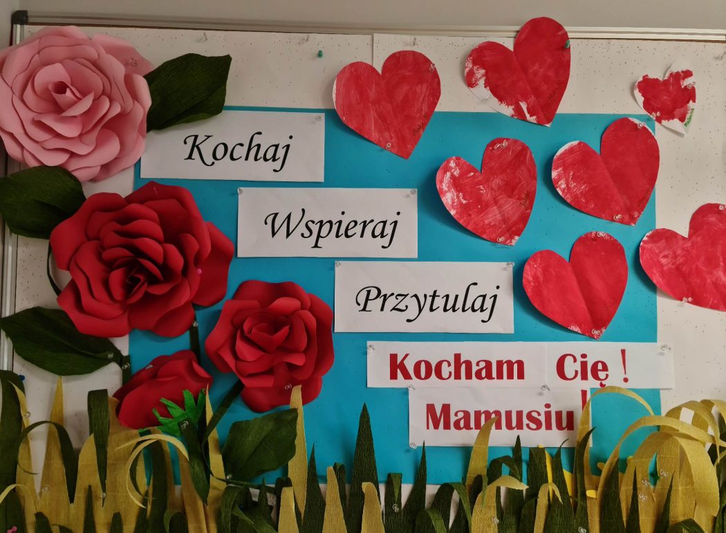 Zdjęcie przedstawia tablice ścienną a na niej dekoracja czerwone róże i serduszka oraz napis "Kochaj, wspieraj, przytulaj- Kocham Cię Mamusiu"
