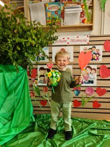 Chłopiec stoi i trzyma bukiecik kwiatów zrobiony z papieru. W tle widać napis "Kocham Cię Mamusiu".