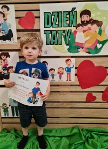 Chłopiec stoi i trzyma dyplom dla Taty. W tle widać zielony napis "Dzień Taty".