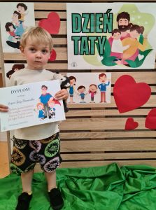 Chłopiec stoi i trzyma dyplom dla Taty. W tle widać zielony napis "Dzień Taty"