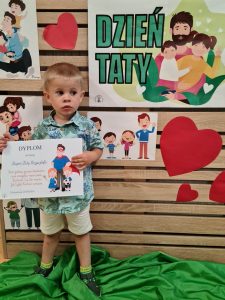 Chłopiec stoi i trzyma dyplom dla Taty. W tle widać zielony napis "Dzień Taty"