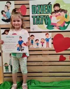 Dziewczynka stoi i trzyma dyplom dla Taty. W tle widać zielony napis "Dzień Taty"