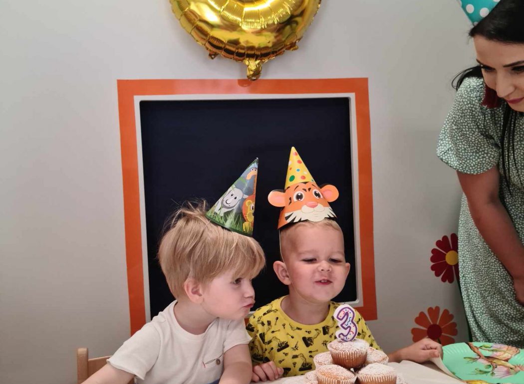 Chłopiec siedzi przy stoliku urodzinowym ubrany w czapeczkę urodzinowąową.