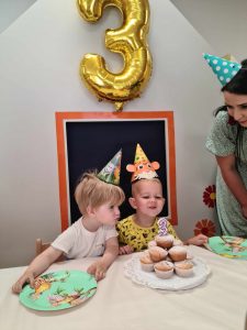 Chłopiec siedzi przy stoliku urodzinowym ubrany w czapeczkę urodzinowąową.