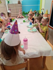Dzieci ubrane w urodzinowe czapki, siedzą przy stoliku i częstują się babeczkami.
