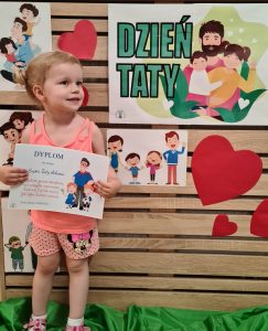 Dziewczynka stoi i trzyma dyplom dla Taty. W tle widać zielony napis "Dzień Taty"