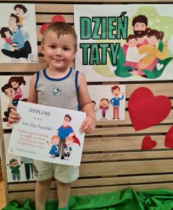 Chłopiec stoi i trzyma dyplom dla Taty. W tle widać zielony napis "Dzień Taty"