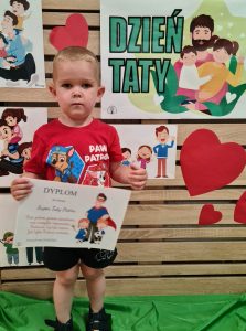 Chłopiec stoi i trzyma dyplom dla Taty. W tle widać zielony napis "Dzień Taty"