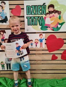 Chłopiec stoi i trzyma dyplom dla Taty. W tle widać zielony napis "Dzień Taty"