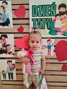 Dziewczynka stoi i trzyma dyplom dla Taty. W tle widać zielony napis "Dzień Taty"