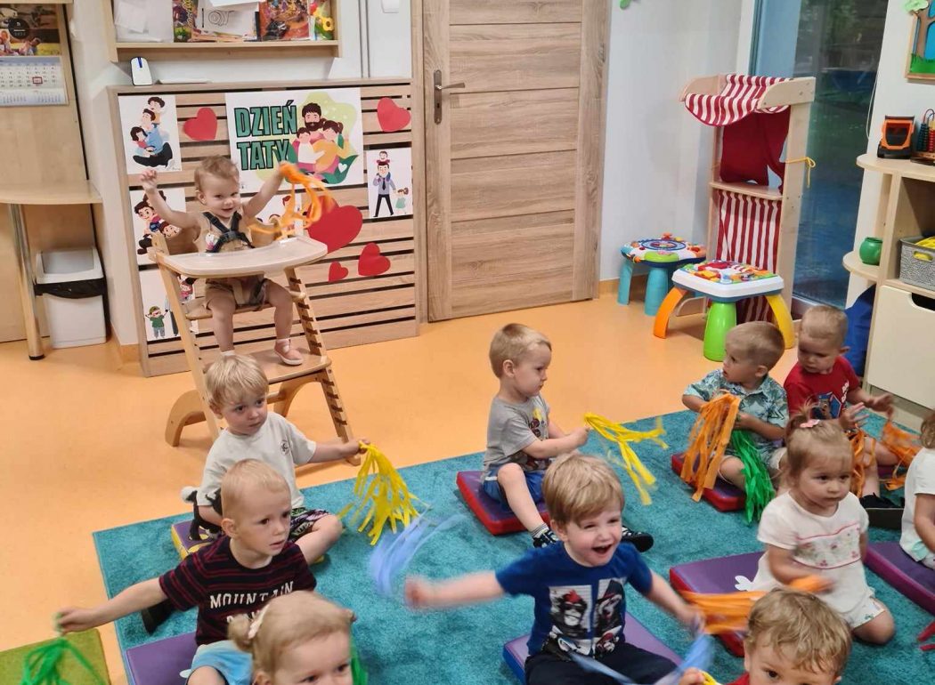 Dzieci siedzą na kolorowych podusiach i trzymają w dłoniach kolorowe pompony. W tle widać sale zabaw.
