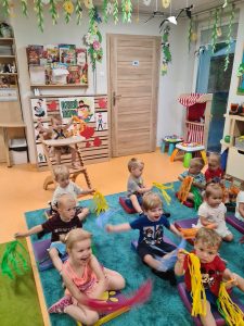 Dzieci siedzą na kolorowych podusiach i trzymają w dłoniach kolorowe pompony. W tle widać sale zabaw.