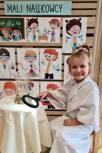 Dziewczynka siedzi ubrana w biały fartuch w dłoni trzyma lupę obok stoi stolik a na nim szklane naczynie z eksperymentem.